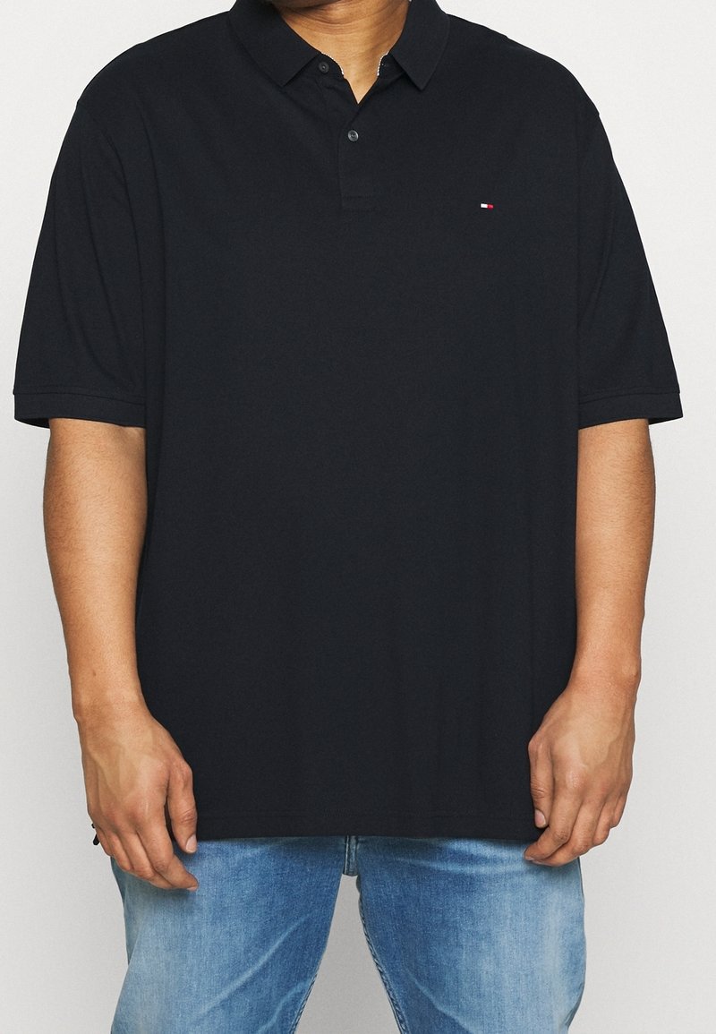 Personne portant un polo noir à manches courtes avec un petit logo sur la poitrine gauche et un jean bleu clair, debout, les mains détendues.