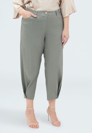 Kobieta ubrana w skrócone, luźne oliwkowo-zielone spodnie z zapinanymi mankietami i beżowe sandały na wysokim obcasie, stojąca z jedną ręką w kieszeni.