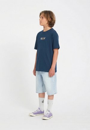 Garçon debout de profil portant un t-shirt graphique bleu marine, un short en denim clair, des chaussettes blanches avec logo, et des baskets violettes sur un fond blanc uni.