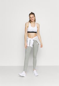 Top sportivo bianco con logo Adidas nero, abbinato a leggins grigi con strisce bianche ai lati. La modella ha accessoriato il look con una camicia bianca annodata.