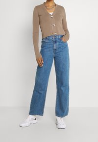 Kvinna iförd en beige knäppt cardigan, avslappnade högmidjade blå jeans, vita sneakers och ett silverhalsband mot en vit bakgrund.