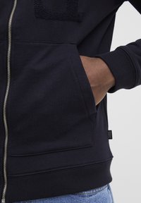 Marineblaue Zip-Hoodie mit strukturiertem, gesticktem Logo auf der Brust, Kängurutaschen und gerippten Bündchen sowie Saum. Glattes Baumwoll-Mischgewebe.