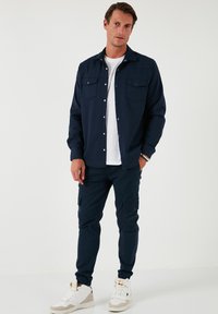 Cămașă închisă la culoare cu nasturi, cu două buzunare pe piept, asociată cu pantaloni cargo tapered și adidași albi. Design relaxat și ajustat, cu o textură netedă.