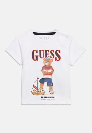 Biała koszulka dla małego dziecka z misiem w stroju marynarza stojącym obok małej żaglówki i napisem „GUESS” dużymi literami nad obrazkiem.
