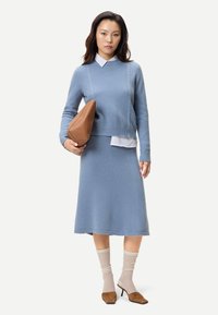 Kvinna står i ljusblå stickad tröja och matchande midikjol, håller en stor brun handväska, beige strumpor och bruna klackskor.