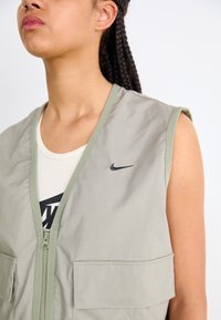 Světle šedá bezrukávová vesta se dvěma předními kapsami, zipovým zapínáním a černým logem Nike na hrudi. Hladká tkanina s matným povrchem.