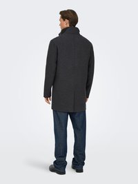 Mann steht mit dem Rücken zur Kamera, trägt einen dunkelgrauen Mantel, blaue Jeans und schwarze Schuhe vor einem schlichten, hellen Hintergrund.