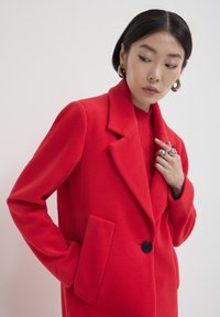 Femme portant un manteau rouge vif et un pull assorti, accessoirisée avec de grandes boucles d'oreilles et de multiples bagues en argent, posant devant un fond uni.