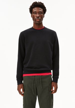 Schwarzer Rundhals-Sweatshirt aus weichem Stoff, mit einem dezenten Logo auf der Brust. Wurde über einem roten Shirt getragen und mit olivgrünen Cargo-Hosen kombiniert.