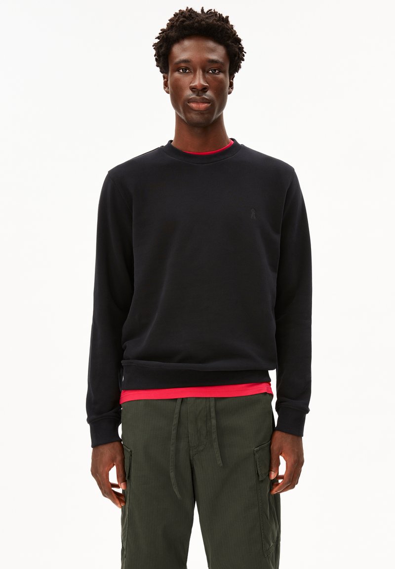 Felpa nera a girocollo in tessuto morbido, con un logo discreto sul petto. Indossata sopra una maglietta rossa e abbinata a pantaloni cargo verdi oliva.