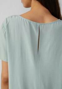 Helles blaus Bluse mit einem Rundhalsausschnitt, kurzen Ärmeln und einem Schlitzdetail im Rücken. Glatte, leichte Stoffqualität mit weicher Textur.