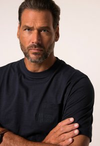 Camiseta de algodón azul marino con escote redondo, mangas cortas y un bolsillo en el pecho con un logotipo de la marca en relieve. Textura suave.