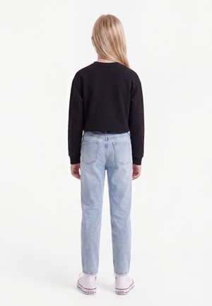 Kind met lang blond haar, draagt een zwarte trui, lichtblauwe spijkerbroek en witte hoge sneakers, staand met de rug naar voren op een witte achtergrond.