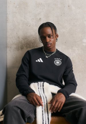 Sort atletisk sweatshirt med hvid Adidas-logo og emblem. Grå jeans og et tekstureret, stribet tæppe draperet over skødet. Betongbaggrund.