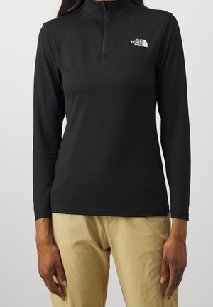 Czarna bluza z długim rękawem zapinana na zamek z logo The North Face na piersi, noszona z beżowymi spodniami, model stojący na jednolitym tle.