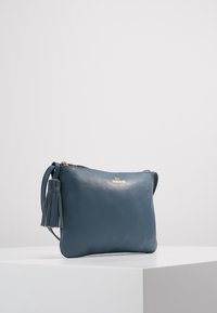 Borsa a tracolla blu in pelle con una texture liscia, caratterizzata da un accentuato ciuffo e un logo dorato sul davanti. Chiusura con zip.
