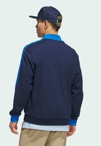Navy sweatshirt with contrasting blue collar and sleeves, featuring a ribbed hem. Model wears beige pants and a navy cap.