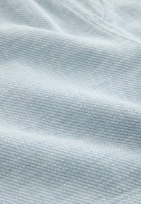 Hellblauer Stoff mit feiner gewebter Textur und sichtbaren Nahtdetails, zeigt subtile Falten und eine weiche Oberfläche.