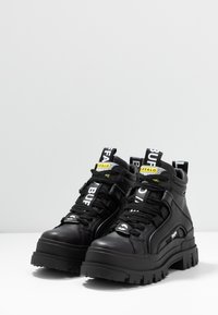 Schwarze High-Top-Sneaker mit einem strukturierten Lederoberteil, dickem Gummisohle, auffälligen Schnürsenkeln und markierten Zuglaschen an der Zunge.