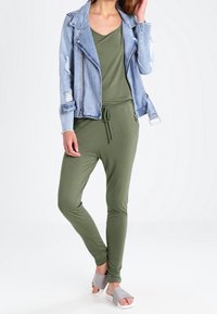 Veste en denim avec des accents usés sur une combinaison verte ; tissu léger, manches longues, taille avec cordon de serrage, associée à des sandales grises.