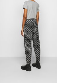 Person som bär svartvitt mönstrade byxor, grå kortärmad skjorta och svarta kraftiga stövlar står mot en enkel vägg.