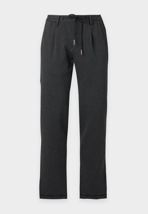 Homme portant une veste noire, un pantalon noir et des bottes noires, tenant un sac de voyage en cuir noir, debout sur un fond blanc uni.