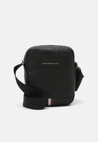 Borsa a tracolla in pelle nera con superficie testurizzata, cinturino regolabile e patch con logo. Accentuata da strisce rosse, bianche e blu nella parte inferiore.