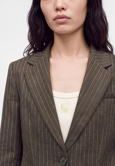 Femme aux longs cheveux foncés portant un blazer marron à fines rayures sur un haut côtelé crème avec un petit emblème brodé.