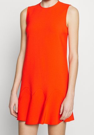 Mujer que lleva un minivestido sin mangas de color naranja brillante con un dobladillo acampanado y con bordes ondulados, de pie frente a un fondo blanco liso.