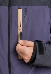 Hand zipping beige zipper on purple and gray Quiksilver jacket with Velcro strap on wrist cuff.