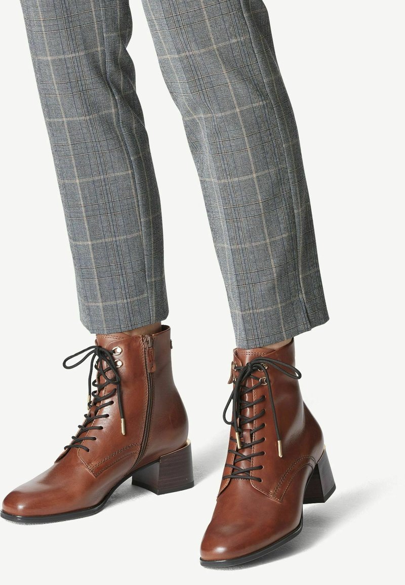 Bottines en cuir marron avec un laçage à l'avant, petit talon et fermeture éclair latérale. Associées à un pantalon tailleur à carreaux gris. Texture lisse.