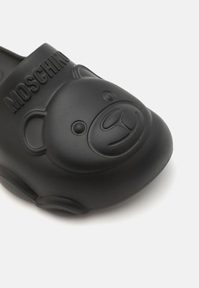 Zueco de goma negra con un diseño de cara de oso de peluche en relieve y logotipo. Textura suave con bordes redondeados y parte trasera abierta para facilitar el uso.