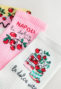 Kolme paari värvilisi sokke: kollased tomatitega, roosad "NAPOLI" tekstiga ja tomatitega, valged pottidega tomatite illustratsioonide ja tekstiga.