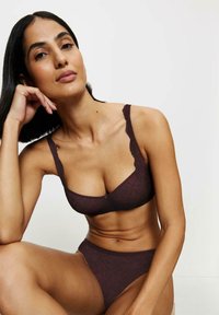 Kobieta o średnim odcieniu skóry i ciemnych włosach, ubrana w ciemnofioletowy koronkowy biustonosz i pasujące majtki, siedzi na jednolitym jasnym tle.