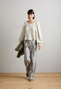 Lichtgroene cropped blouse met textuurafwerking, vloeiende mouwen; grijze geplooide broek; olijfgroene jas over de arm; neutrale achtergrond.