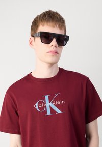 Crne pravokutne sunčane naočale s tamnim lećama, nošene preko bordo majice s bijelim i svjetloplavim logotipom Calvina Kleina.