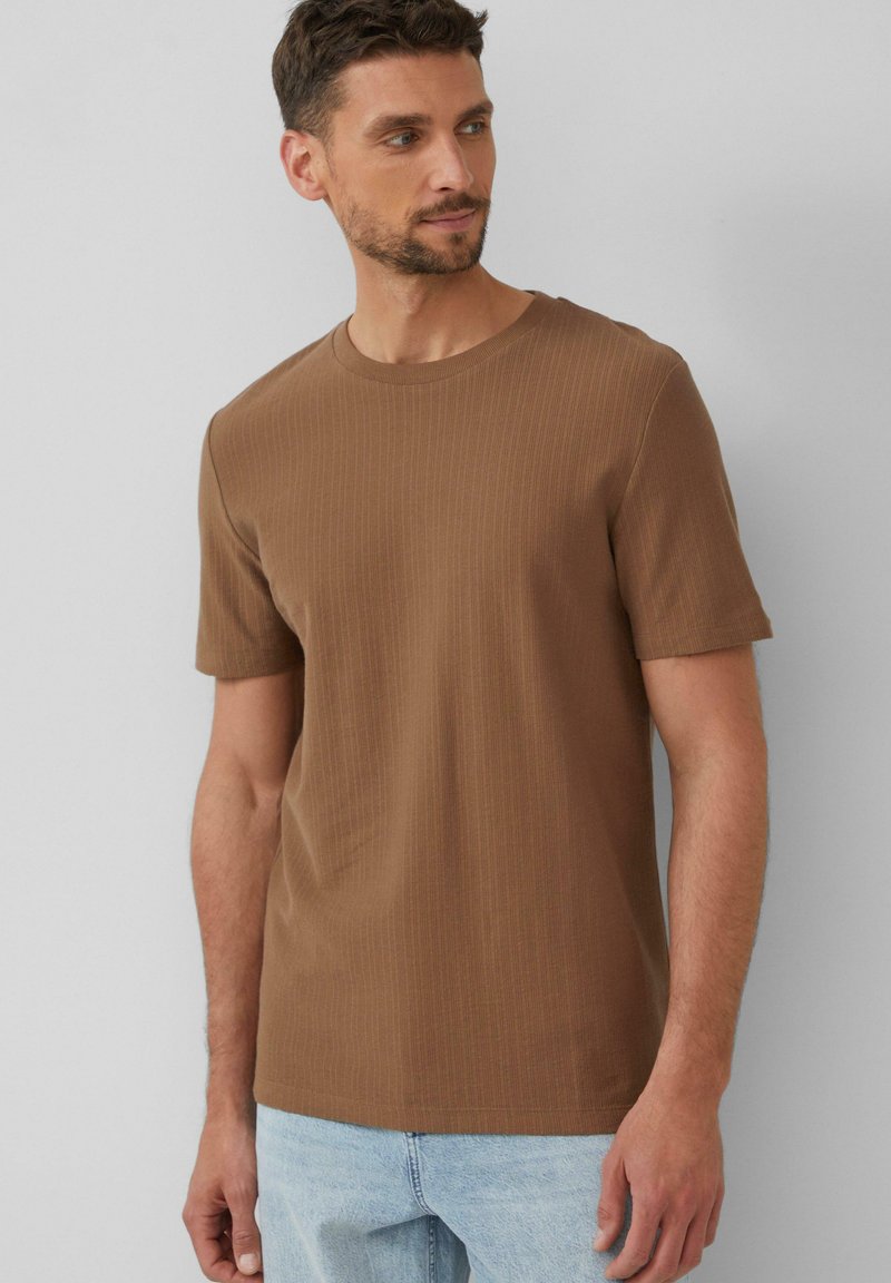 Braunes geripptes T-Shirt mit Rundhalsausschnitt und kurzen Ärmeln. Der Stoff wirkt weich mit einer dezenten Textur und wird mit hellblauen Jeans kombiniert.