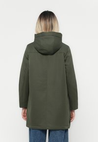Parka imperméable vert avec capuche, présentant une coupe droite, des manches longues et une texture lisse. Présentée de dos.