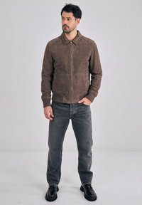 Mies seisoo päällään ruskea mokkatakki, tummanharmaat farkut ja mustat kengät vaaleanharmaan yksivärisen taustan edessä.