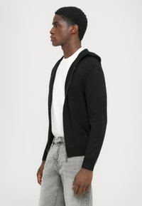 Sort zip-up hoodie lavet af strikket stof; har en frontlynlås, lange ærmer og ribbede manchetter. Bæres over en hvid T-shirt og grå jeans.