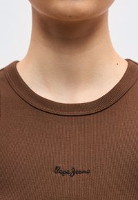 Κοντινό πλάνο του λαιμού και του άνω μέρους του στήθους ενός ατόμου που φοράει καφέ ριγωτό πουκάμισο με την επιγραφή "Pepe Jeans" κεντημένη σε μαύρο στο στήθος.