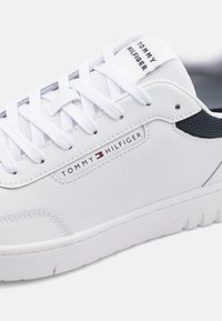 Sneaker en cuir blanc avec une finition texturée, fermeture à lacets et accents navy. Présente un détail de logo sur le côté et une semelle en caoutchouc.