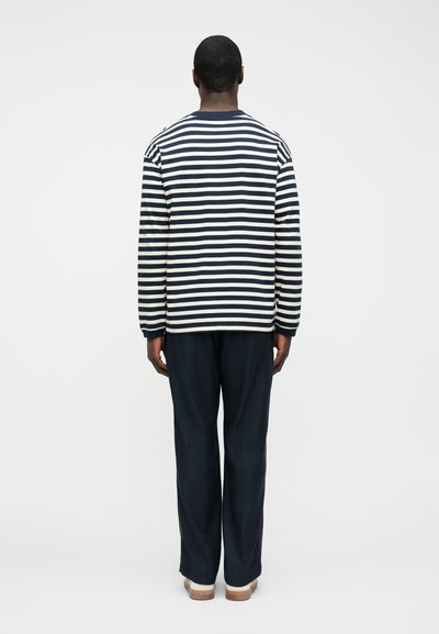 Άνδρας με την πλάτη προς τα έξω, φοράει μπλε ναυτικό και λευκό ριγέ μπλουζάκι με μακριά μανίκια, σκούρο παντελόνι και μπεζ παπούτσια, σε απλό φόντο.