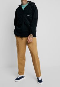 Black pull-over jacket with a hood, two front pockets, and adjustable drawstring. Paired with tan pants and navy sneakers.