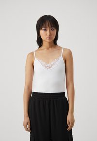 White camisole with a v-neck, featuring a lace trim along the neckline. Worn with loose black pants. Smooth fabric, thin adjustable straps.