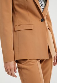 Žena v camel hnědém přizpůsobeném blejzru a odpovídajících kalhotách s černým knoflíkem, pod kterým je vidět bílá vzorovaná blůza.