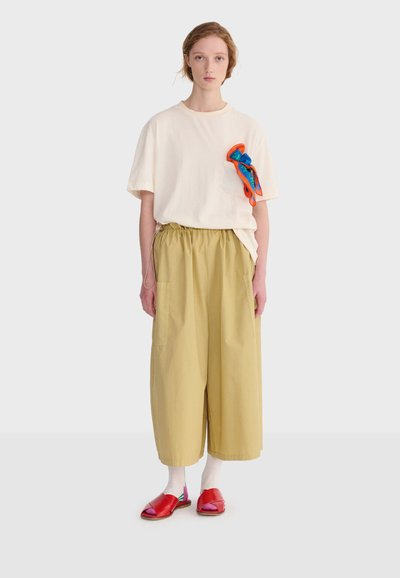 Jeune personne portant un T-shirt oversize crème avec une pochette orange-bleue, un pantalon large beige ample, des chaussettes blanches et des sandales rouges ouvertes.