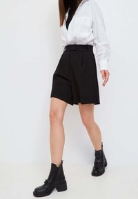 Pantalones cortos negros ajustados con pliegues, cintura alta, combinados con una camisa blanca de gran tamaño y botas negras de tobillo. Tejido suave y ajuste estructurado.