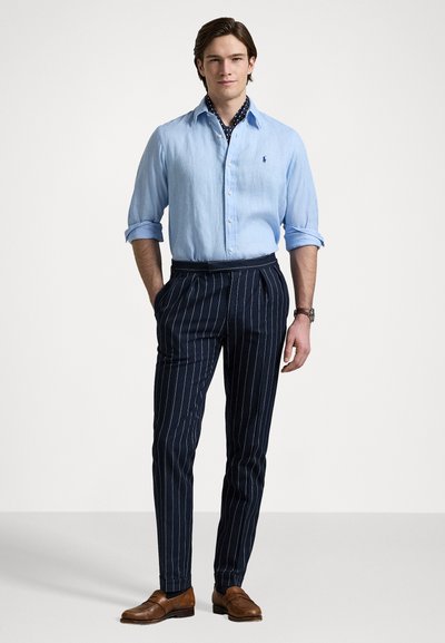 Világoskék gombos ing, sötét tengerészkék csíkos nadrág, barna bőr cipők, és a mintás gallér részlet látható az ing alatt.