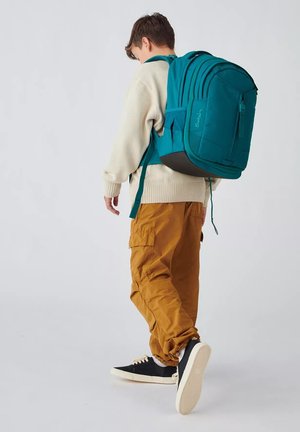 Mochila de color turquesa con correas acolchadas y bolsillos laterales. La lleva puesta una figura con un suéter marfil y pantalones cargo mostaza, con zapatillas negras.